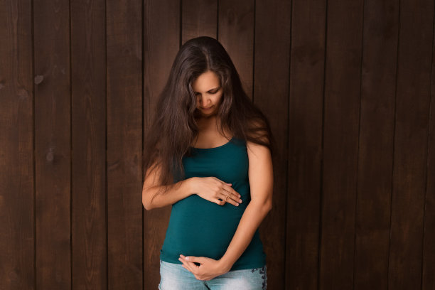 身着牛仔裤短裤和绿色上衣的孕妇双手搭在深棕色的腹部上。怀孕、分娩、准备和期望概念。美丽柔情的怀孕照片图片下载