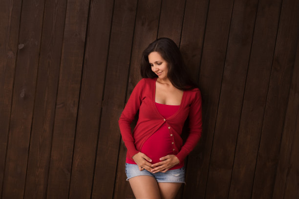 圣诞节孕妇在牛仔裤短裤和红色上衣牵手腹部在一个深棕色的背景。怀孕，母性，期望概念。美丽柔情的怀孕照片图片下载