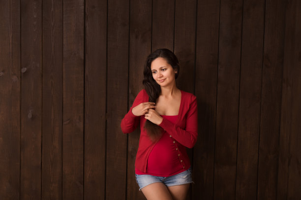 圣诞节孕妇在牛仔裤短裤和红色上衣牵手腹部在一个深棕色的背景。怀孕，母性，期望概念。美丽柔情的怀孕照片图片下载