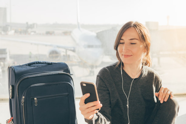 一名年轻女子带着她的手提行李和智能手机在机场候机厅等待她的航班(刚过了免税期)。丢失或取消航班-需要赔偿概念(巴塞罗那，西班牙)。图片下载