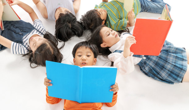 孩子们躺在白色的地板上看书，头挨着头，围成一圈，幼儿园图书馆，幼儿园教育理念图片下载