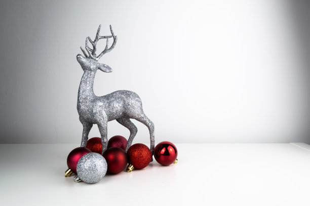 闪闪发光的驯鹿桌面圣诞装饰品矗立在白色的反光表面上，周围是闪亮的红色和银色的圣诞树装饰品。图片下载
