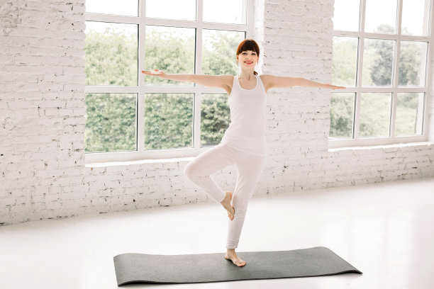 年轻的运动女性在家里练习瑜伽，站在Vrksasana锻炼，树姿势，工作，穿着白色运动服，室内全长，窗户背景图片下载