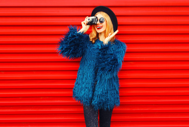 穿着蓝色人造皮草大衣，戴着圆帽和太阳镜，在彩色的红色墙壁背景下，用复古相机拍摄照片图片下载