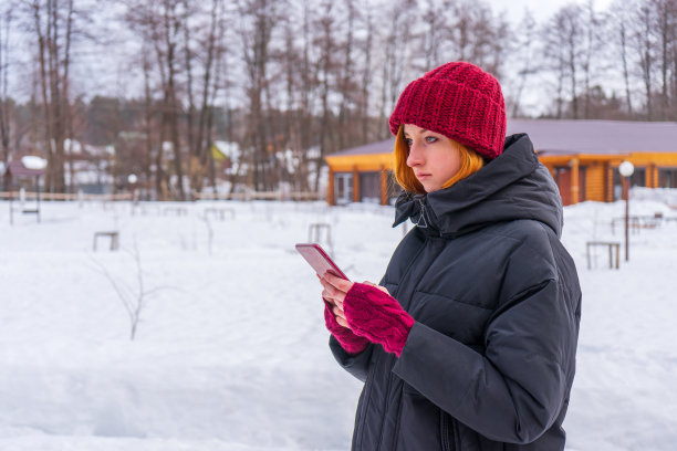 年轻可爱的女孩穿着黑色羽绒服，戴着酒红色的帽子和无指手套，手拿智能手机站在户外，面对冬天的乡村景观。周末在乡村，青少年的时尚概念图片下载