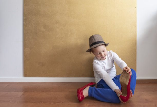 孩子，一个四岁的金发女孩，留着短发，戴着帽子。一个穿着白色t恤和蓝色裤子的女孩坐在地板上，试穿红色的女鞋。一个孩子在一堵橙色墙的背景下模仿妈妈。图片下载