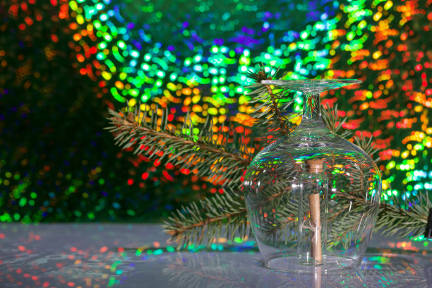 一张折成管状的纸条，用线系起来。上面盖着一个葡萄酒杯。以冷杉树枝和彩虹为背景的散景。图片下载