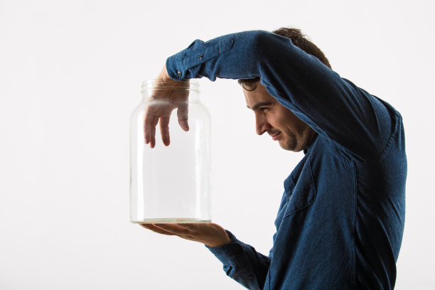 一个好奇的商人拿着一个空玻璃罐试图把他的手放在里面看鬼鬼祟祟的眼睛孤立在白色的背景。图片下载