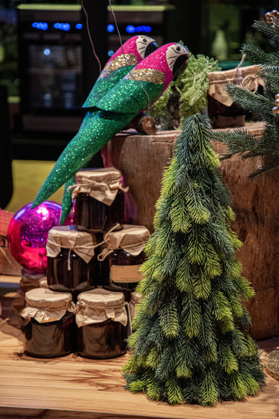 圣诞装饰品。两只闪闪发光的玩具鹦鹉站在树桩上，金字塔形的玻璃罐子里放着自制的果酱。人工绿色圣诞树前景。图片下载