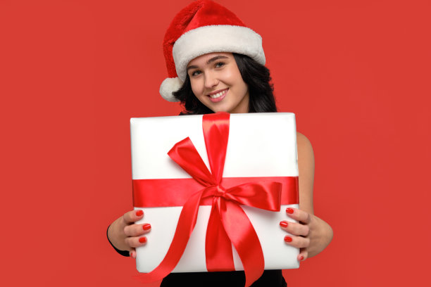 女人穿着一件连衣裙和圣诞老人的帽子微笑着拿着圣诞礼物盒白色的红丝带图片下载