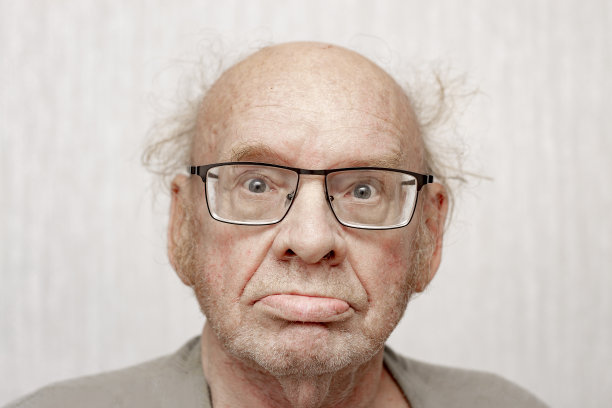 一位没有发型、穿着灰色衣服的老人正看着照相机。特写肖像。几何尺寸。图片下载