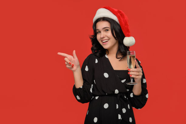 黑发女孩穿着黑色连衣裙，戴着圣诞老人的帽子，拿着一杯香槟，指着抄写空间图片下载