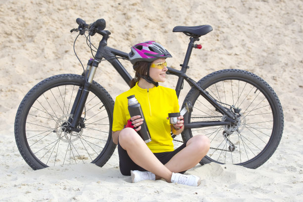 美丽的女孩骑着黄色的自行车，手里拿着茶和保温瓶，背景是沙子。体育和娱乐。自然和人类图片下载