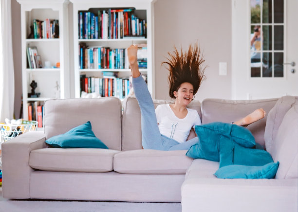 快乐的少女有乐趣在家里跳和跌落在客厅的沙发上。休闲的周末活动图片下载