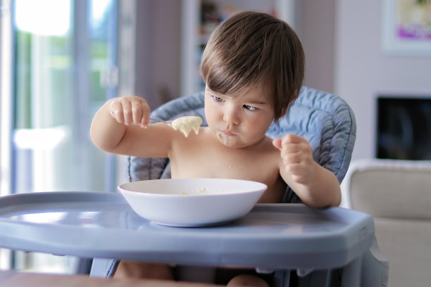 有趣的小男孩吃粥看着满勺在他的手坐在高椅子。孩子学会自己吃饭。图片下载