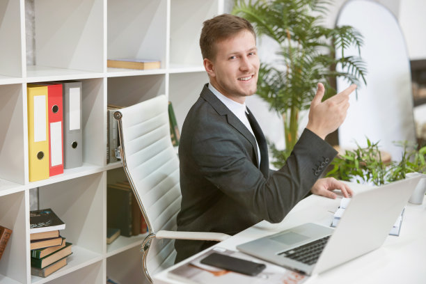 微笑英俊的男人经理在他的笔记本电脑工作，坐在他的工作地点在办公室看起来很开心。多任务、工作概念图片下载