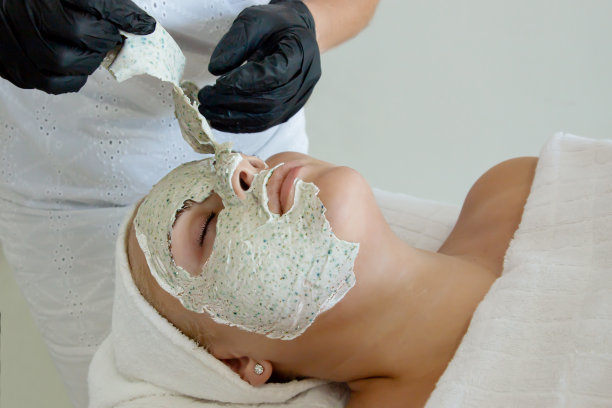 面部皮肤治疗。美容师戴着黑手套从病人脸上取下口罩。在美容院接受美容手术的妇女图片下载