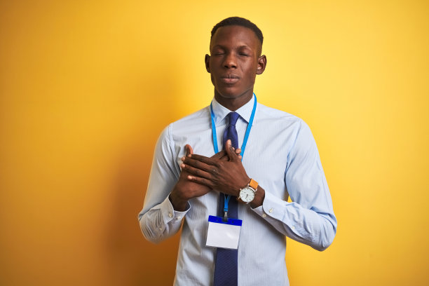 非洲裔美国商人戴着身份证在孤立的黄色背景微笑，双手放在胸前，闭着眼睛，脸上有感激的手势。健康的概念。图片下载