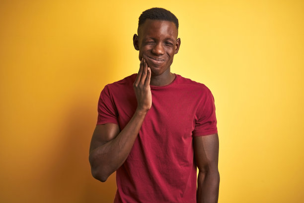 年轻的非洲裔美国人身穿红色t恤站在孤立的黄色背景与手触摸嘴痛苦的表情，因为牙痛或牙齿疾病的牙齿。牙医的概念。图片下载