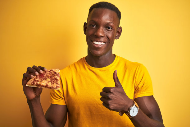 非洲裔美国人吃比萨饼站在孤立的黄色背景高兴地大微笑做ok的手势，拇指与手指竖起，优秀的手势图片下载