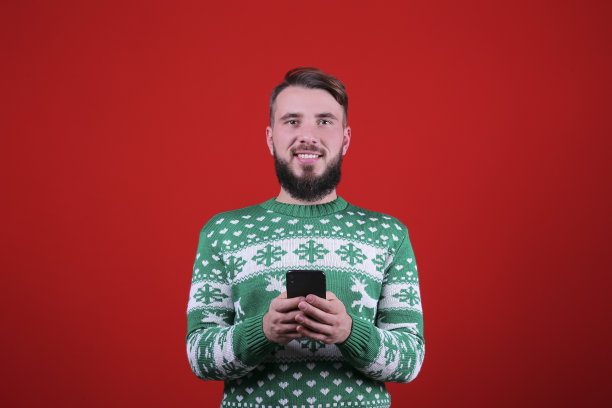 红墙背景上，一个留着完美胡须的年轻人穿着圣诞服装。图片下载