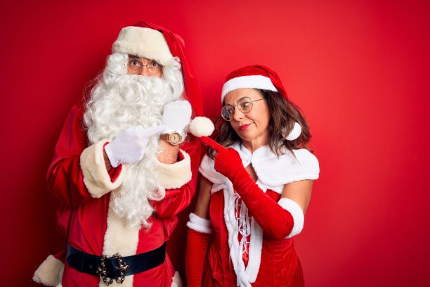 一对中年夫妇穿着圣诞老人的服装，戴着眼镜越过孤立的红色背景，匆忙地指着手表时间，不耐烦地看着镜头，用轻松的表情图片下载