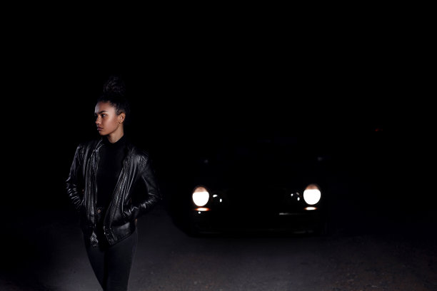 一个穿着皮夹克和黑色衣服的年轻女孩在一条废弃的沙路上摆姿势。晚上在灯光下的汽车前灯和灯笼图片下载