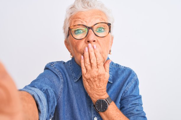 灰白头发的女人穿着牛仔衬衫和眼镜在孤立的白色背景下自拍，用手捂着嘴因错误而震惊，表达恐惧，在沉默中害怕，秘密的概念图片下载