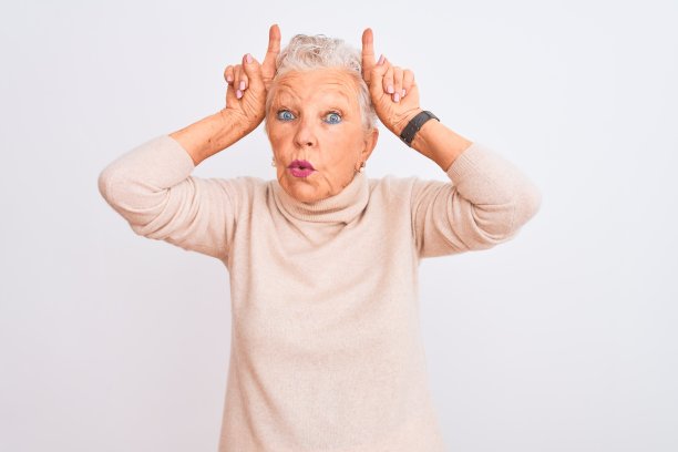 年长的灰白头发的女人穿着高领毛衣站在孤立的白色背景做有趣的手势，手指在头上像牛角图片下载