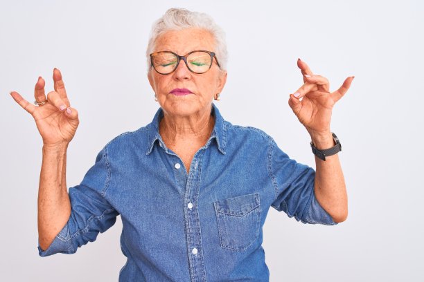 年长的灰白头发的女人穿着牛仔衬衫和眼镜在孤立的白色背景放松，微笑着闭上眼睛做冥想手势与手指。瑜伽的概念。图片下载