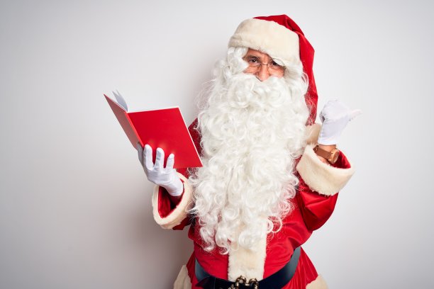 中年男子穿着圣诞老人服装在孤立的白色背景下看书，用手和大拇指向上指着背后，自信地微笑着图片下载