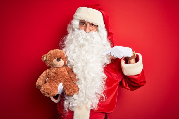 中年男子穿着圣诞老人服装，抱着泰迪熊在孤立的红色背景与惊讶的脸指向自己的手指图片下载