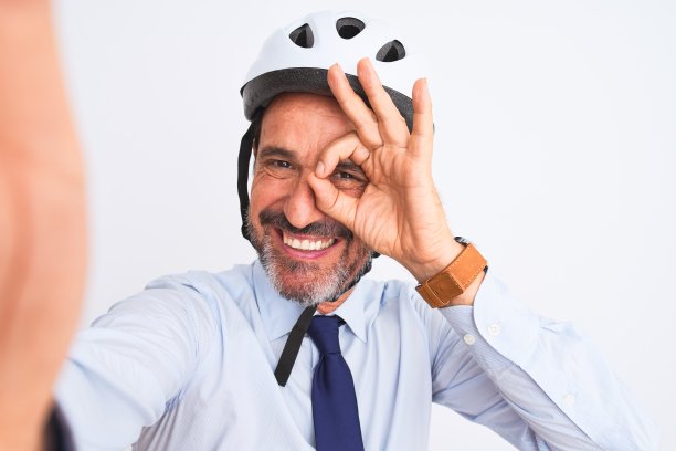 中年商人戴着自行车头盔，在孤立的白色背景与快乐的脸微笑做ok手势与手在眼睛透过手指看图片下载