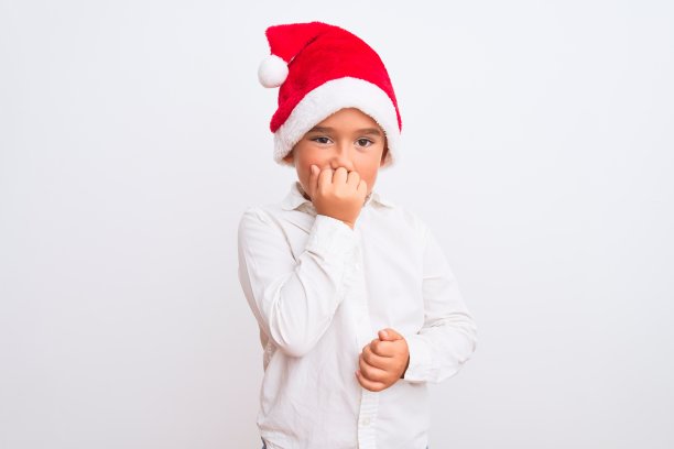 漂亮的男孩戴着圣诞老人的帽子站在孤立的白色背景看起来紧张和紧张的手在嘴咬指甲。焦虑的问题。图片下载