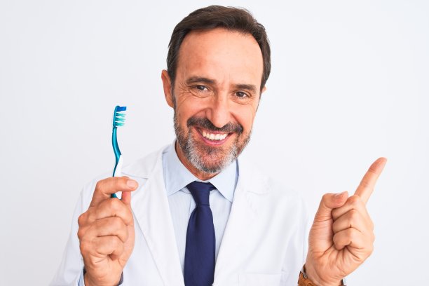 中年牙医拿着牙刷站在孤立的白色背景非常高兴地用手和手指指着一边图片下载