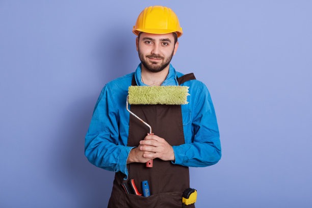 图片的胡子英俊的男子穿着蓝色制服，黄色保护头盔和棕色围裙，吸引人的画家手中拿着他的工具，准备油漆与油漆辊。建筑的概念。图片下载