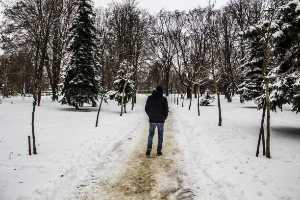 一个穿着黑色夹克的人正在路上走。一个男人的剪影。冬天的路。冬天戴兜帽的男人。黑色的衣服和牛仔裤。一个有树和雪的冬日。图片下载