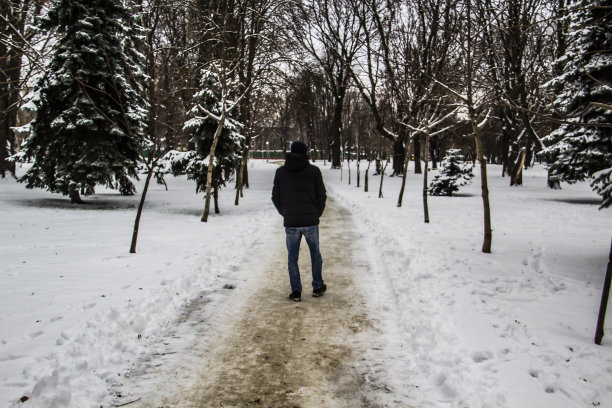一个穿着黑色夹克的人正在路上走。一个男人的剪影。冬天的路。冬天戴兜帽的男人。黑色的衣服和牛仔裤。一个有树和雪的冬日。图片下载