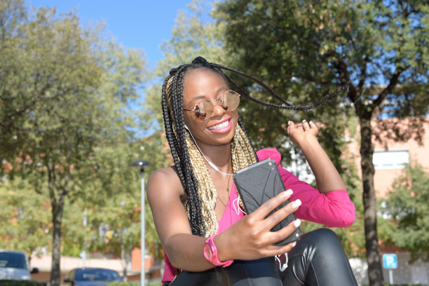 快乐的非洲风格的黑人女孩与长黑和金发辫子拍照时，微笑着用她的耳机。她在一个小镇的大街上图片下载