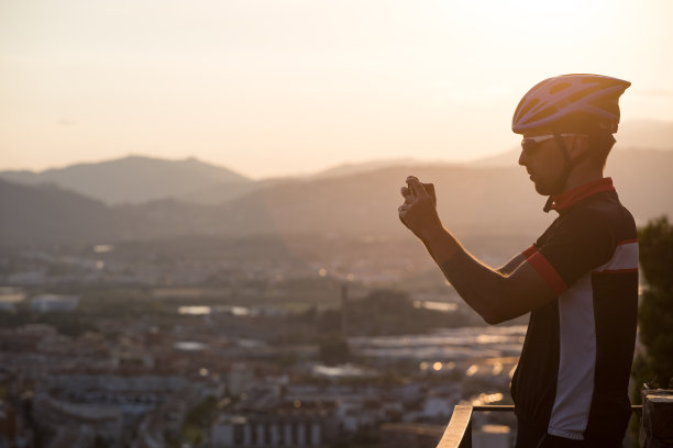 骑车者用智能手机拍照。旅行者在智能手机上拍摄照片，在手机上使用手机，旅游博客。在西班牙的一座山上，一个骑着自行车的男人用手机拍下了日落时分的照片图片下载