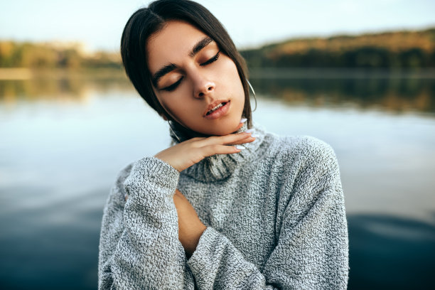 特写年轻的黑发女子闭着眼睛享受温暖的天气，穿着灰色毛衣，摆姿势在自然背景。湖光背景上摆姿势的美女，有着梦幻般的表情。图片下载