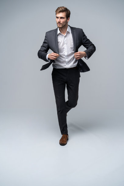 对每一步都充满信心。一身西装的英俊青年在灰色的背景下一边调整他的夹克一边走在前面。图片下载