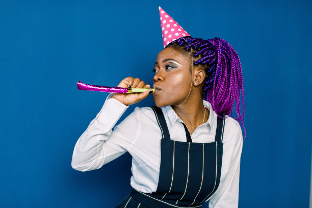 生日派对，新年狂欢。年轻微笑的非洲妇女在蓝色背景庆祝明亮的事件，穿着优雅时尚的白色裙子和黑色裤子，与粉红色的派对帽与噪音。图片下载