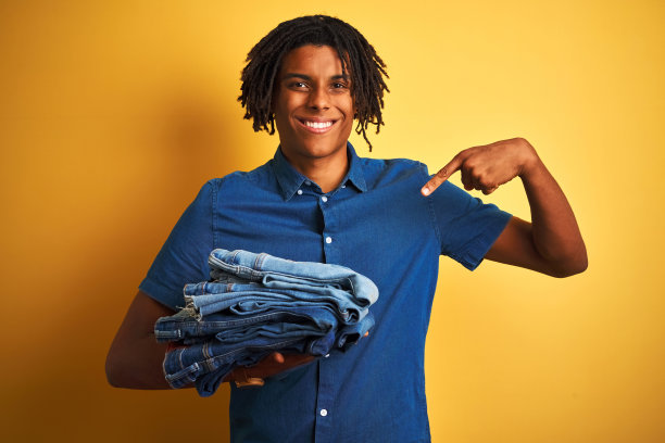 留着雷鬼发的非洲人拿着一叠叠叠的牛仔裤盖在孤立的黄色背景上，惊讶地用手指着自己的脸图片下载