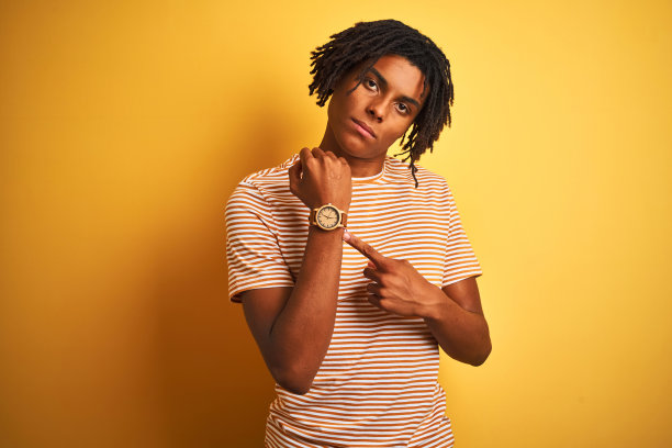 一个留着雷鬼辫、穿着条纹t恤的黑人站在孤立的黄色背景上，急匆匆地指着手表时间，不耐烦地看着镜头，用轻松的表情图片下载