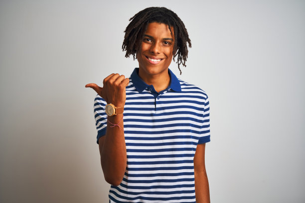 非洲人与雷鬼发穿着条纹蓝色马球站在孤立的白色背景微笑与快乐的脸看着和指向一边拇指向上。图片下载