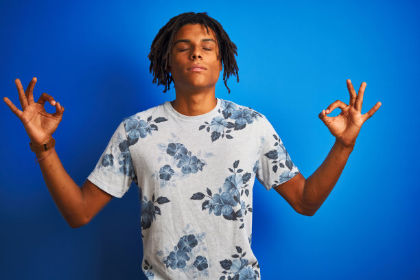 美国黑人男子与雷鬼发穿着夏天花t恤在孤立的蓝色背景放松和微笑闭上眼睛做冥想手势与手指。瑜伽的概念。图片下载