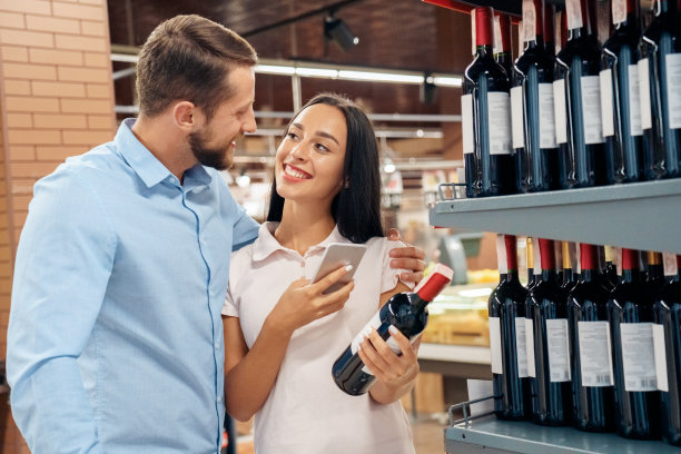 日常购物。一对情侣在超市的酒类部拥抱选择酒女浏览智能手机看着对方开心图片下载