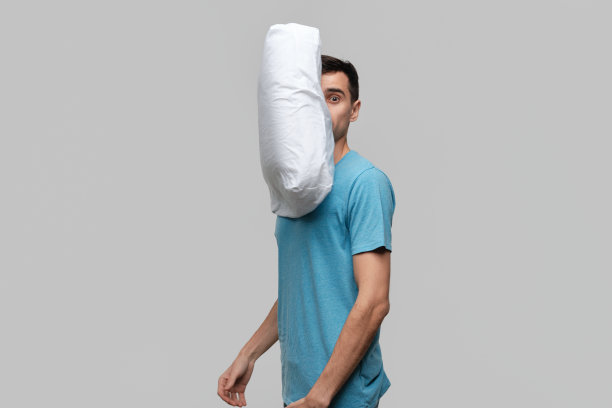 一个穿着蓝色t恤的漂亮的深色头发男子被白色枕头飞向他的方向震惊了。图片下载