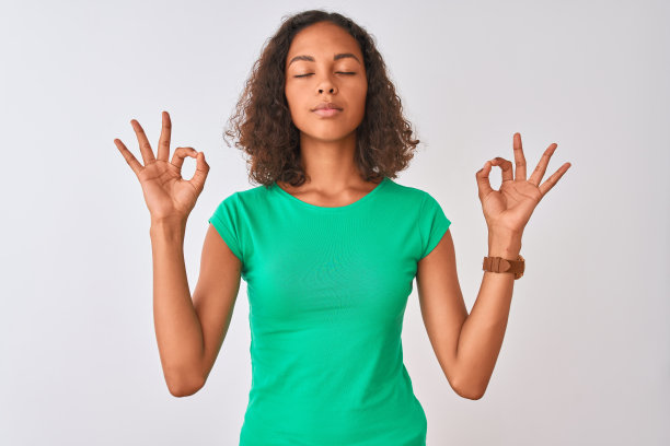 年轻的巴西女子穿着绿色t恤站在孤立的白色背景放松和微笑，闭上眼睛做冥想手势与手指。瑜伽的概念。图片下载